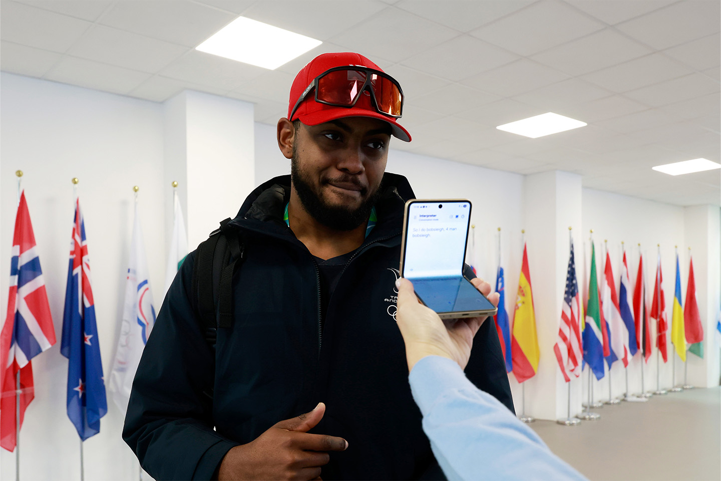 Xaverri Williams se encuentra frente a una serie de banderas nacionales, frente a la pantalla de portada de un Galaxy Z Flip7 Olympic Edition en manos de otro atleta. La pantalla principal presenta la interfaz del Intérprete.