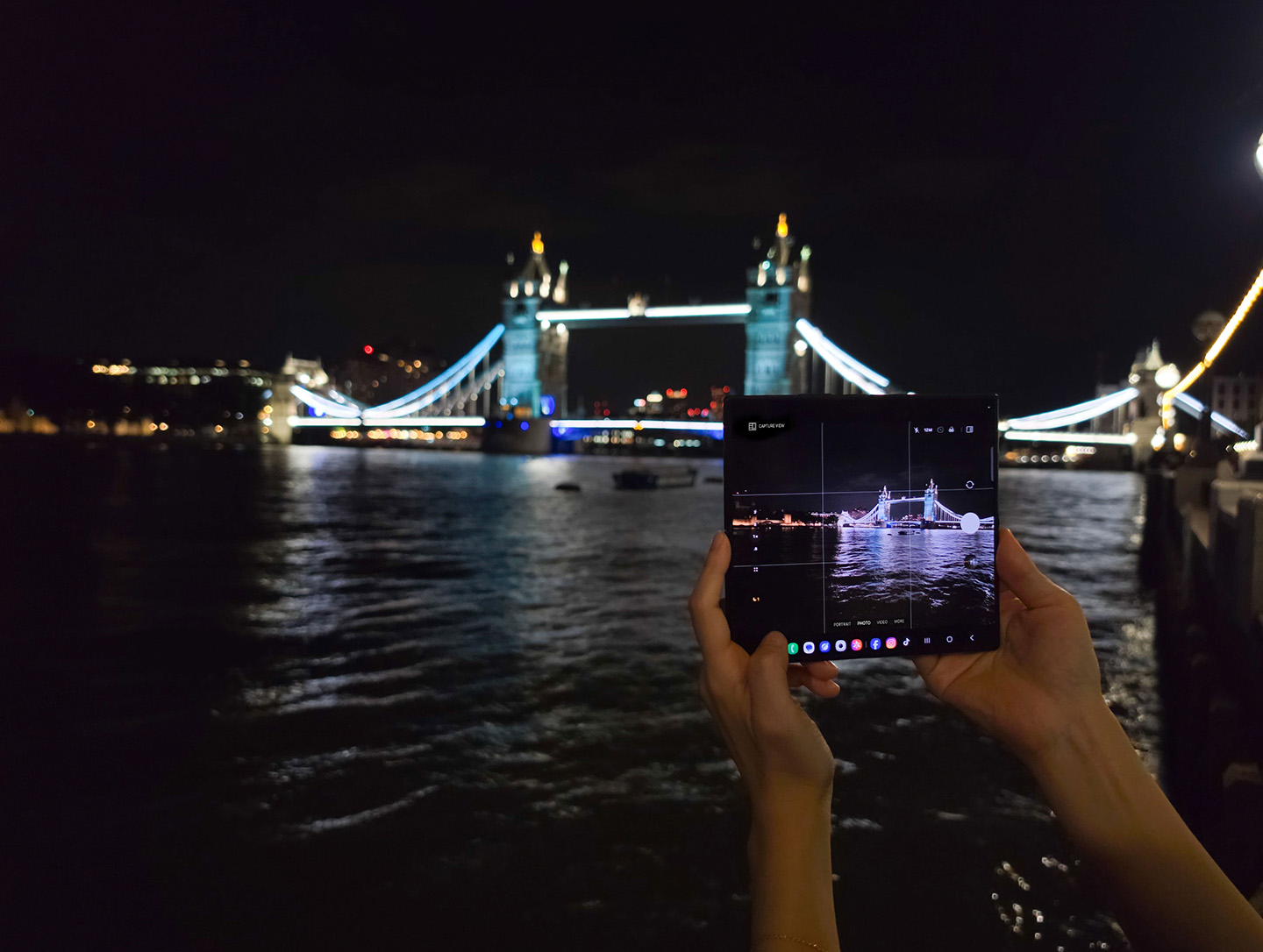 Hands holding a Galaxy Z Fold8 Ultra displaying Tower Bridge on its screen at night, with the illuminated bridge and Thames River visible in the background under moonlight in London. 
