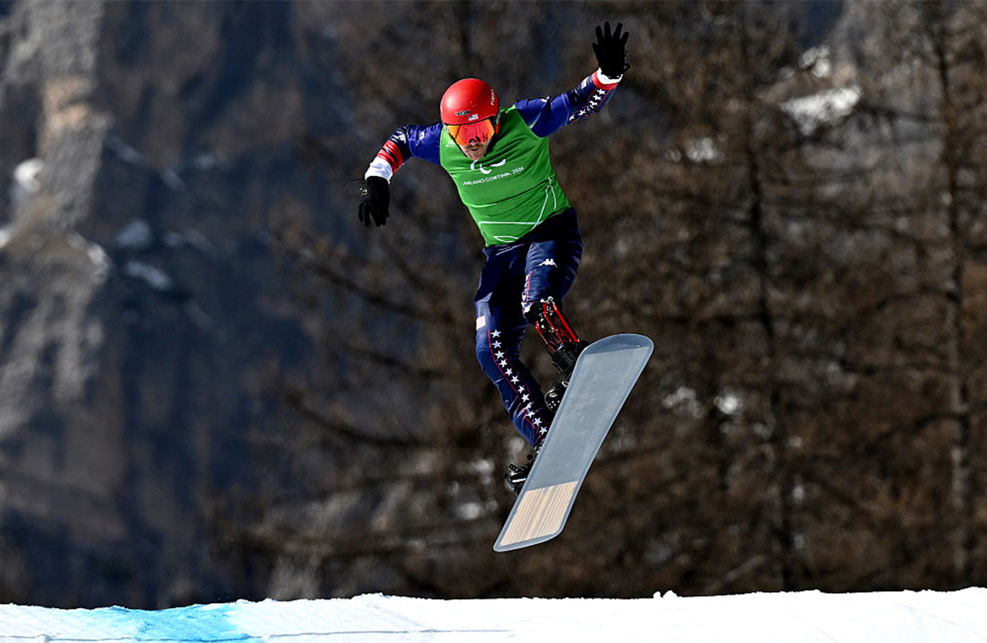 A photo of Paralympian Mike Schultz on a snowboard, airborne, jumping over some snow with trees in the background.