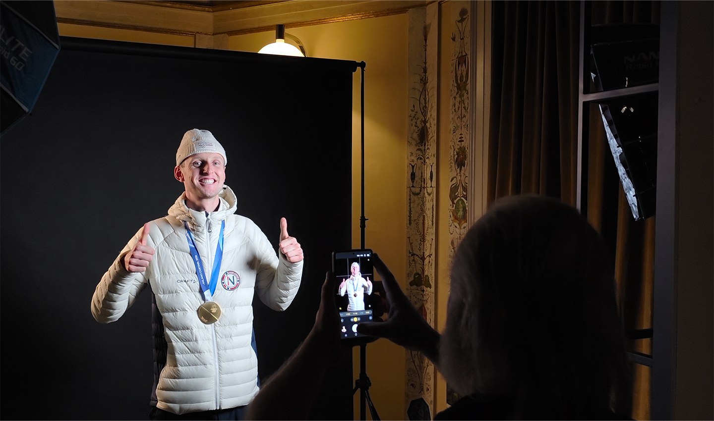 An Olympic athlete wearing Norway’s official Olympic hat and jacket, and his Olympic gold medal, poses for a photo in front of a black screen with a big smile and two thumbs up.