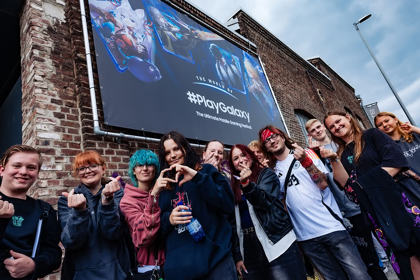 A crowd of people stand in front of the event building featuring The World of PlayGalaxy banner, making heart signs with their hands towards the camera.