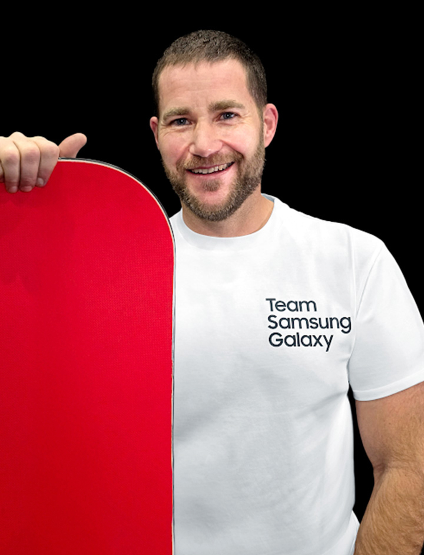 A photo of Paralympian Mike Schultz wearing a white Team Samsung Galaxy t-shirt and holding a red snowboard in his right hand.