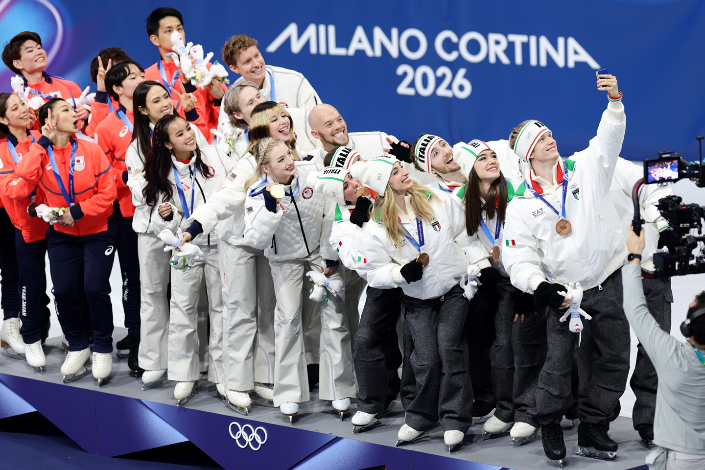 19 Olympic athletes stand in a rough line on a stage and look right to pose together for a selfie being taken by the rightmost using a Galaxy Z Flip7 Olympic Edition.