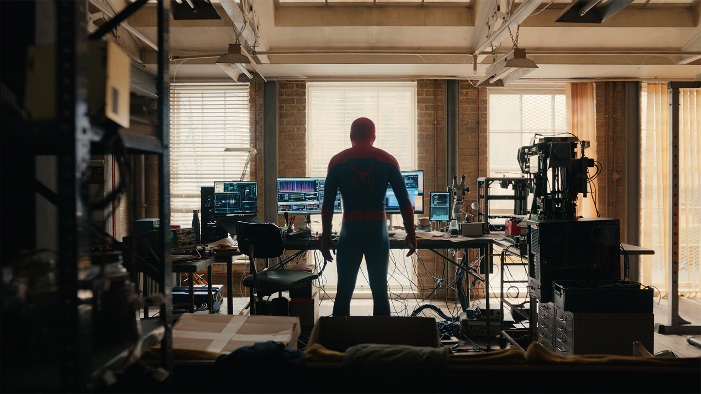 Spider-Man stands with his back facing the viewer, in front of a desk covered with various tech devices.