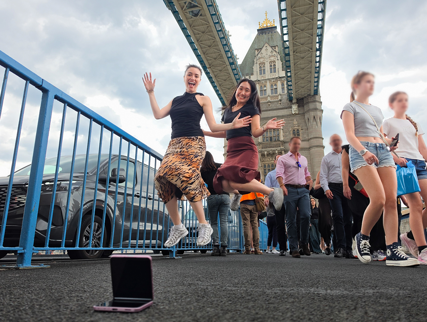 Galaxy Z Flip8 in Flex Mode propped on ground at Tower Bridge with two women jumping and posing for a hands-free selfie with the bridge and sky visible in background. 