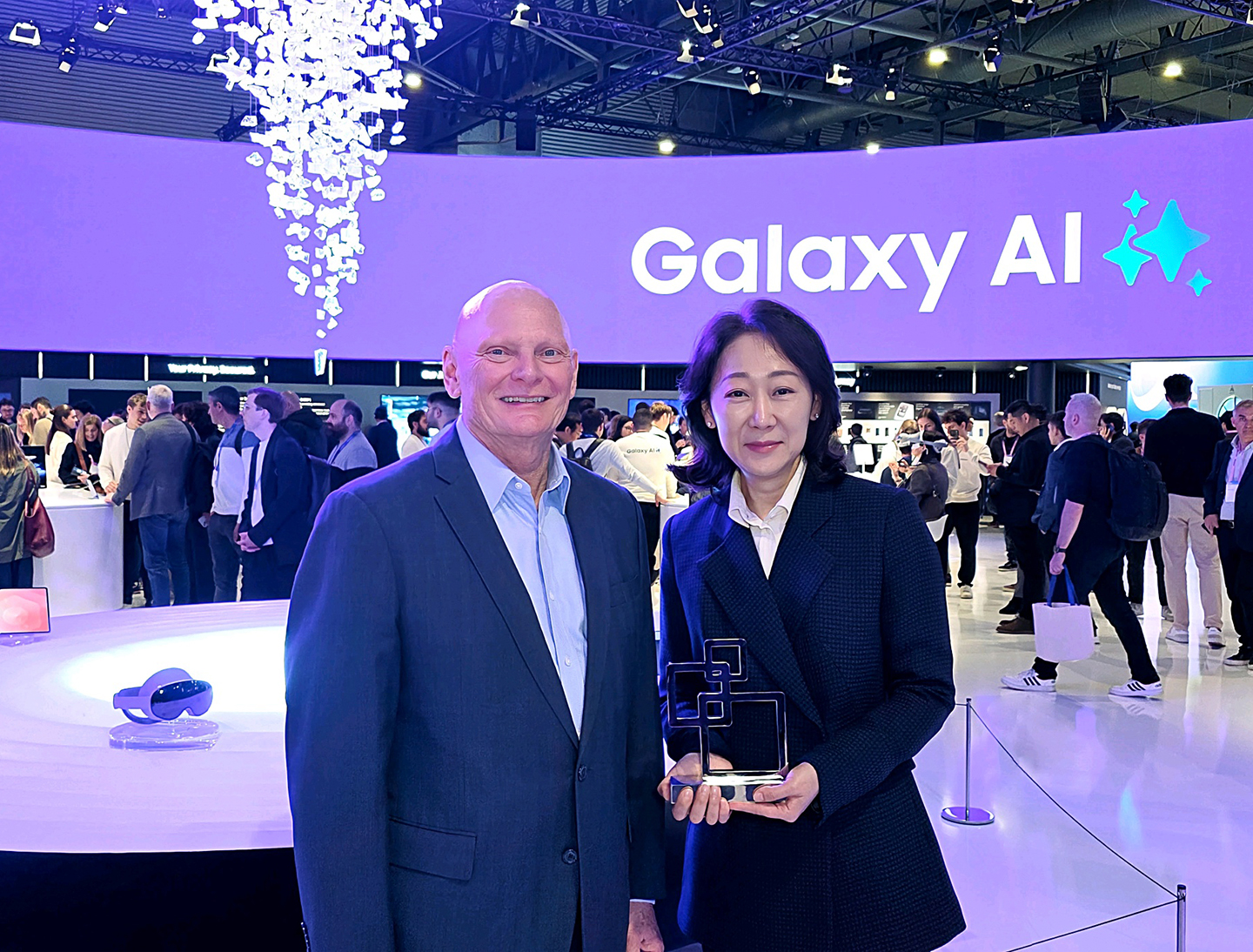 Samsung EVP Stephanie Choi holds the Best in Show award while standing next to GSMA CEO John Hoffman in a busy exhibition hall at MWC 2026.