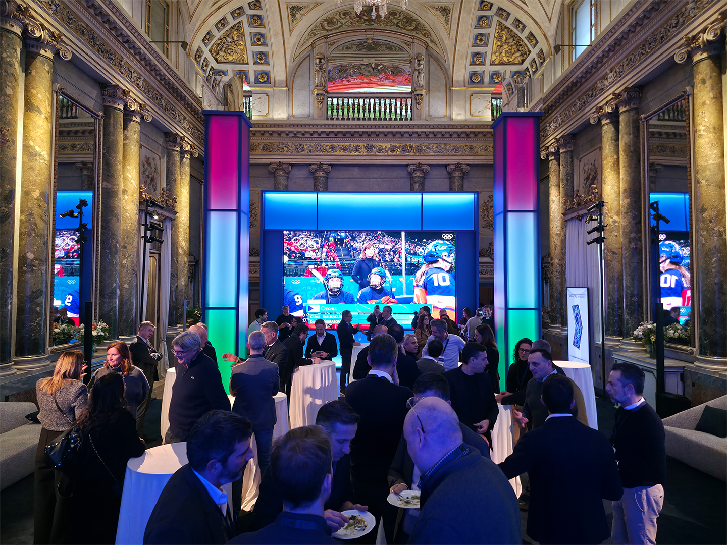 Many people talk and mingle in a room with a high-vaulted ceiling, gold inlaid columns and a large display screen on the far wall showing Olympic footage.