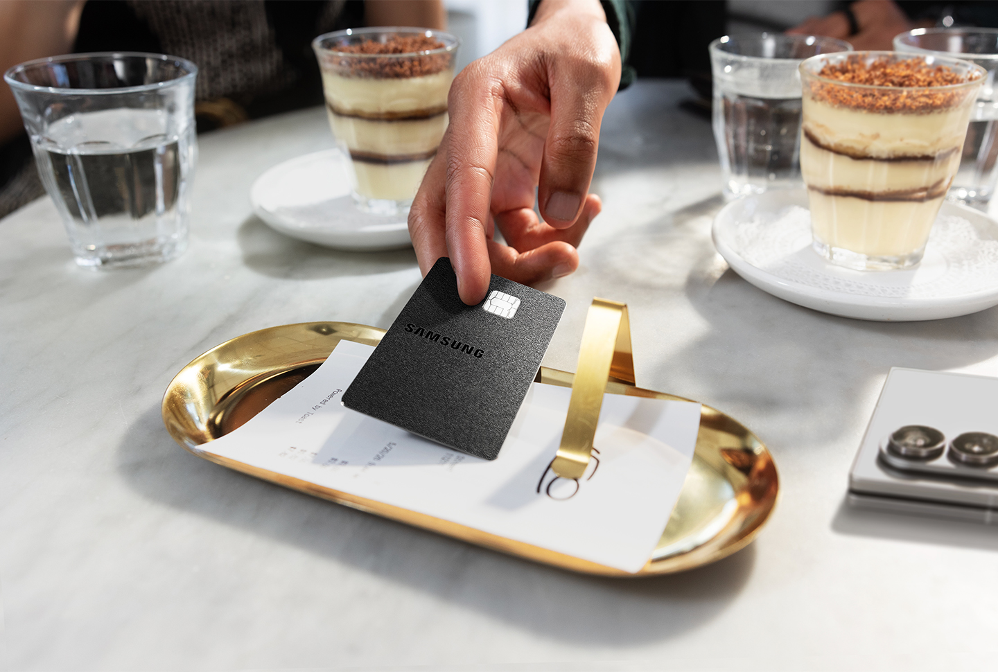 A hand places the Samsung Galaxy Card onto a golden tray on a table with two desserts and several glasses of water in the background.